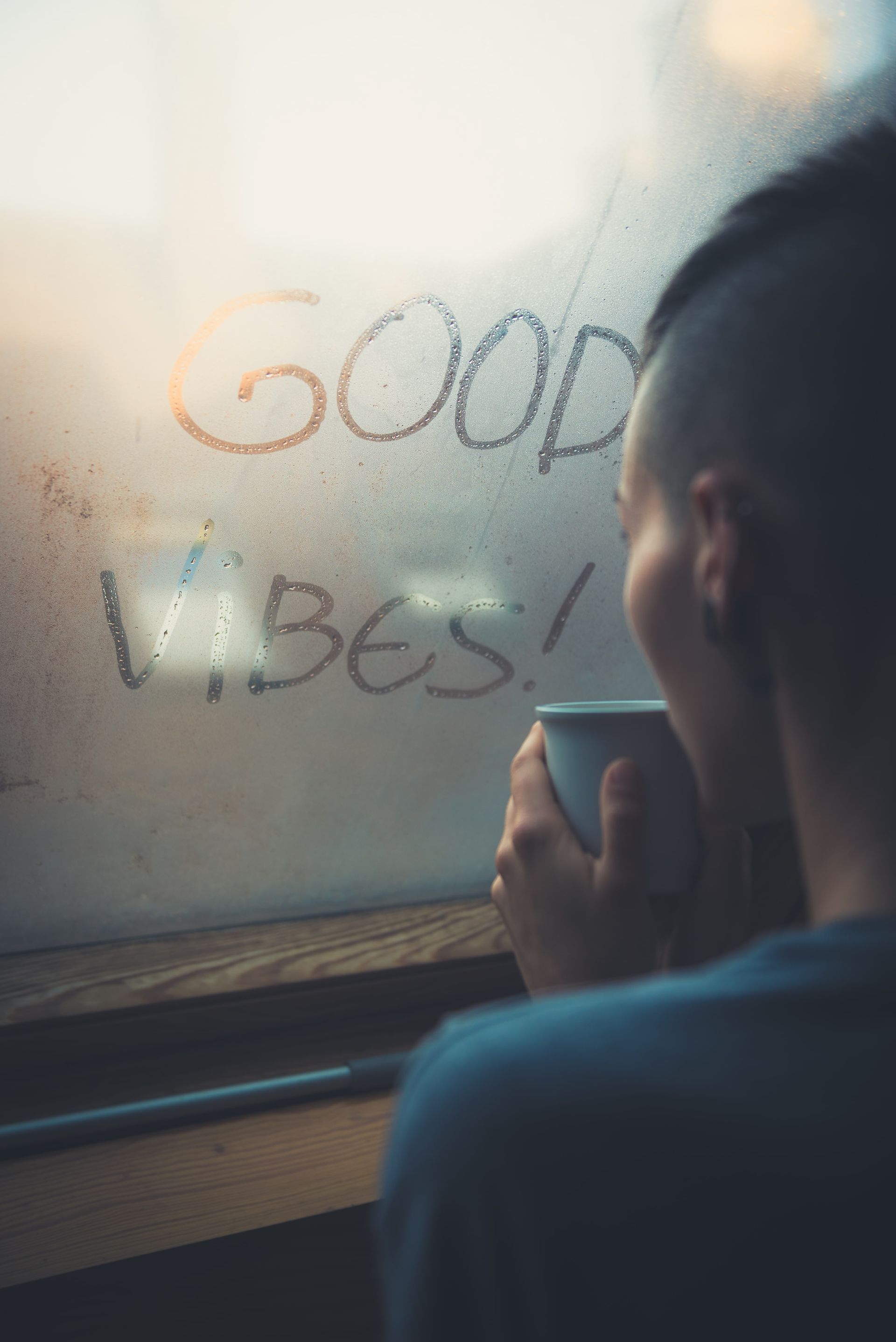A man is holding a cup of coffee in front of a window with the words `` good vibes '' written on it.