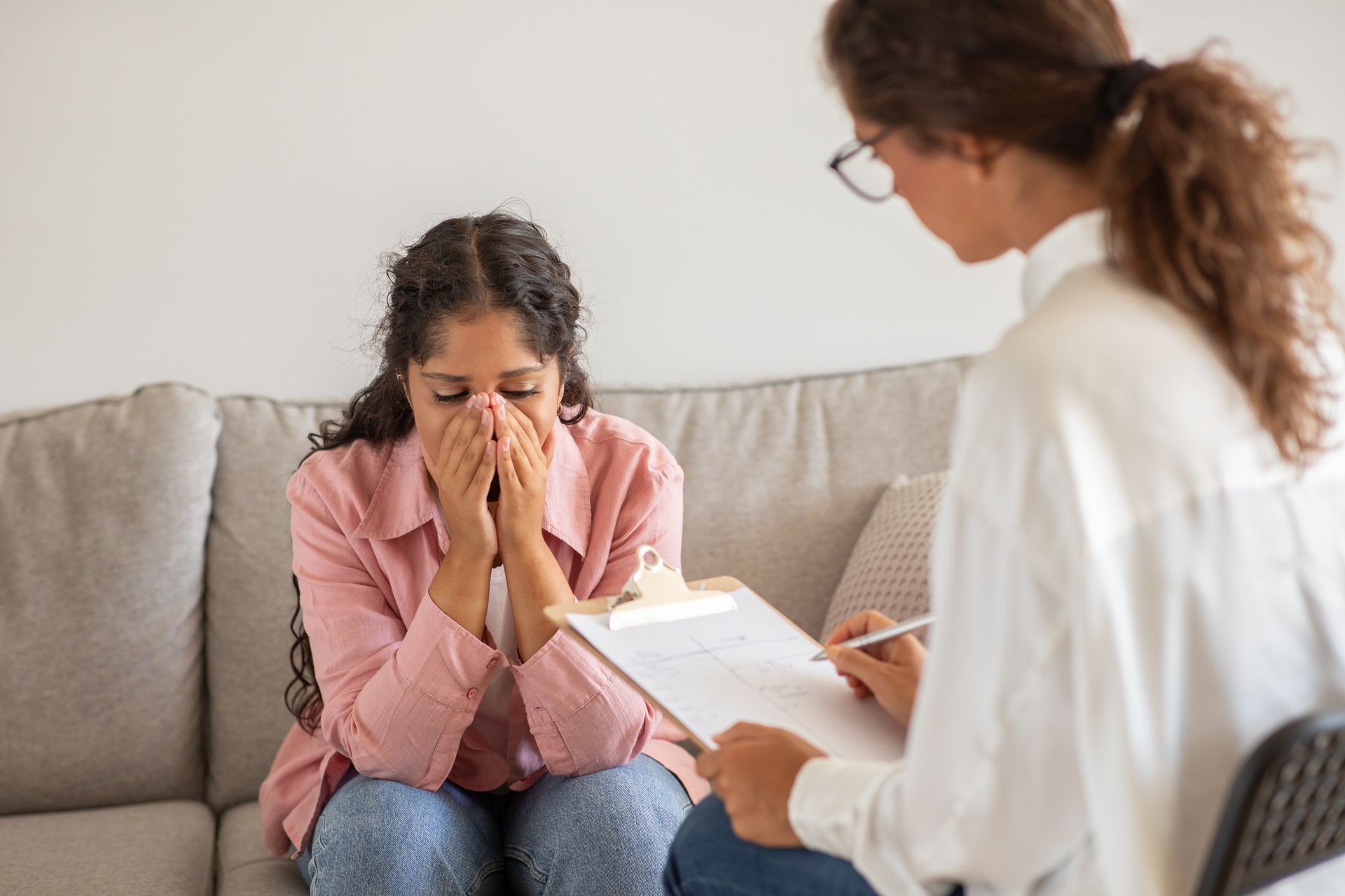 A woman is sitting on a couch covering her face while talking to a doctor.