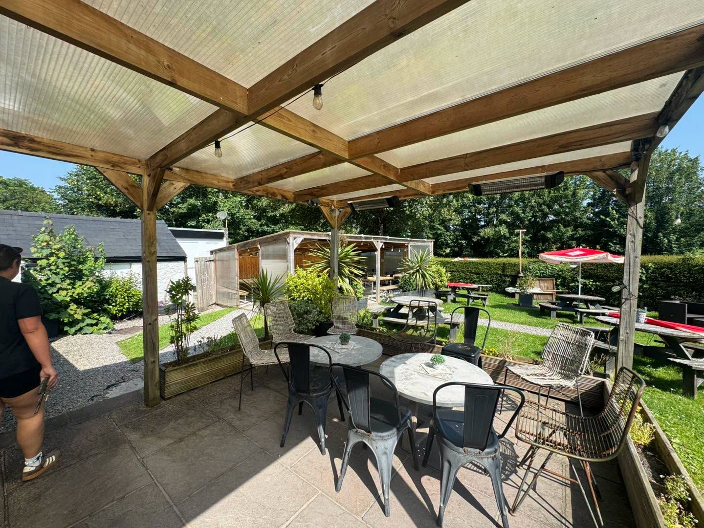 The Bush Inn, St Hilary | Covered patio with table & chairs, overlooking a sunny garden with picnic benches & red umbrellas.