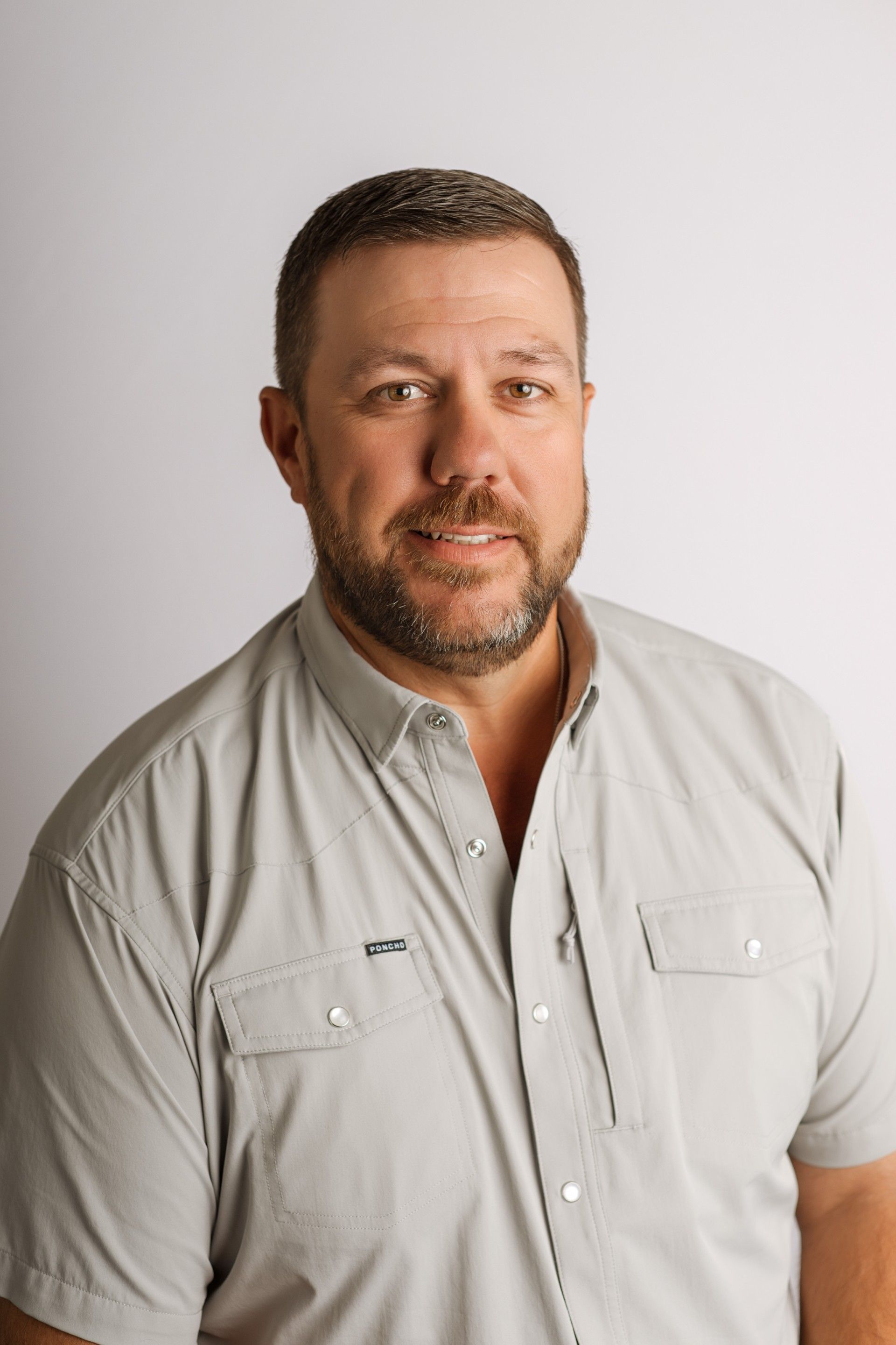 Man with beard and short hair in a light gray button-up shirt smiling at the camera. White backdrop.