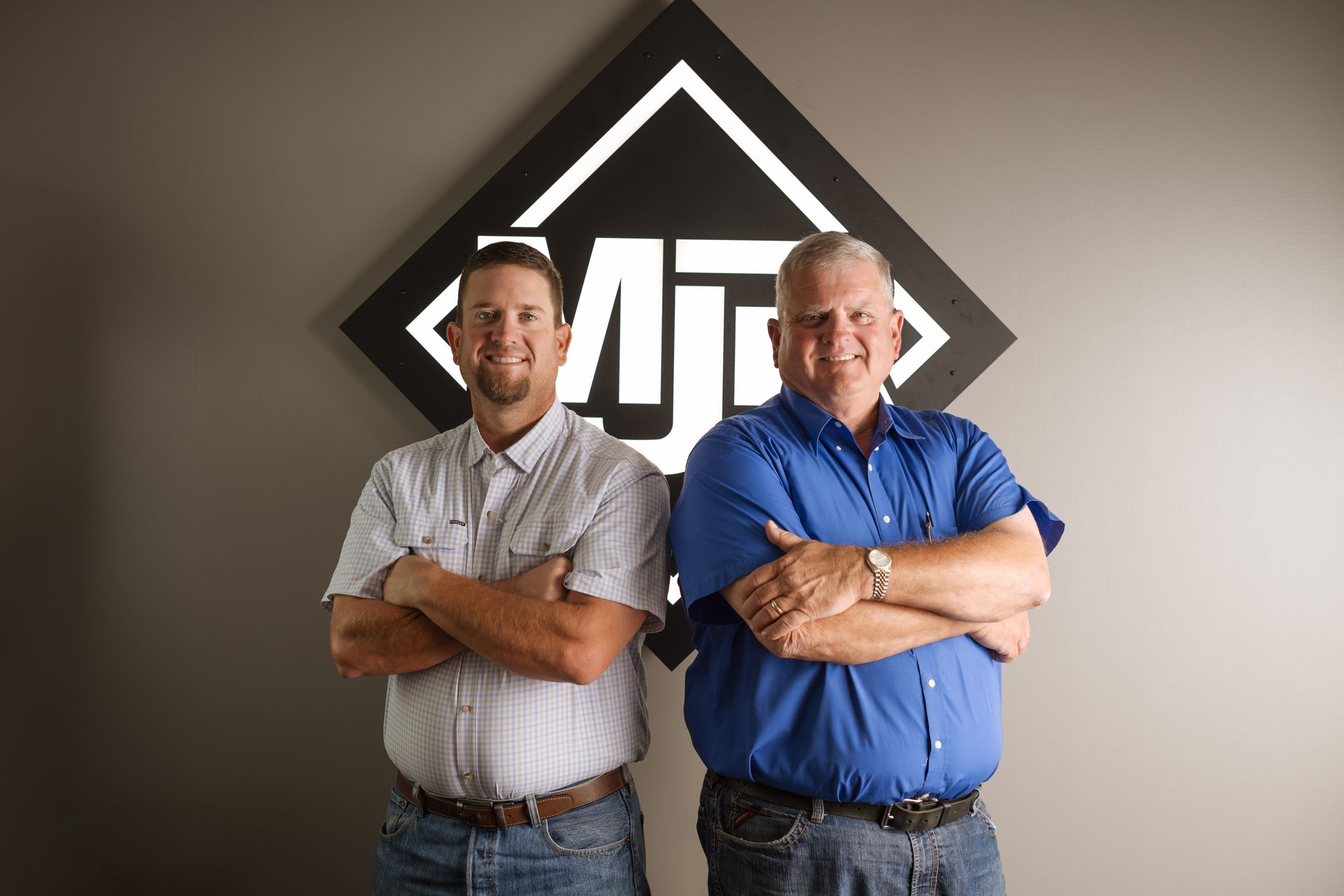 Two men with arms crossed in front of a logo. One in a blue shirt, the other in a button-down.