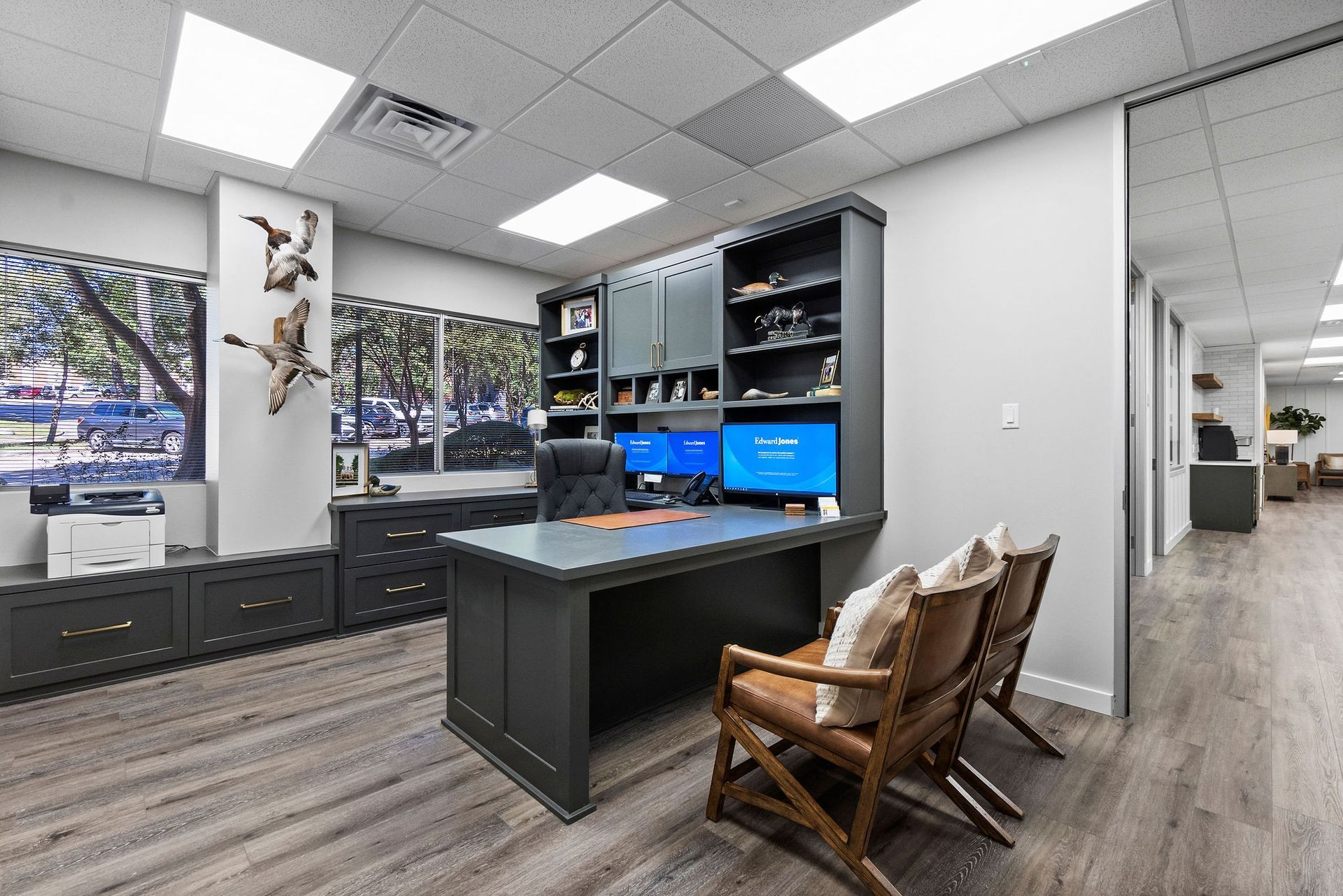 Office with dark gray desk and built-in storage. Two monitors, leather chairs, and taxidermied ducks are visible.