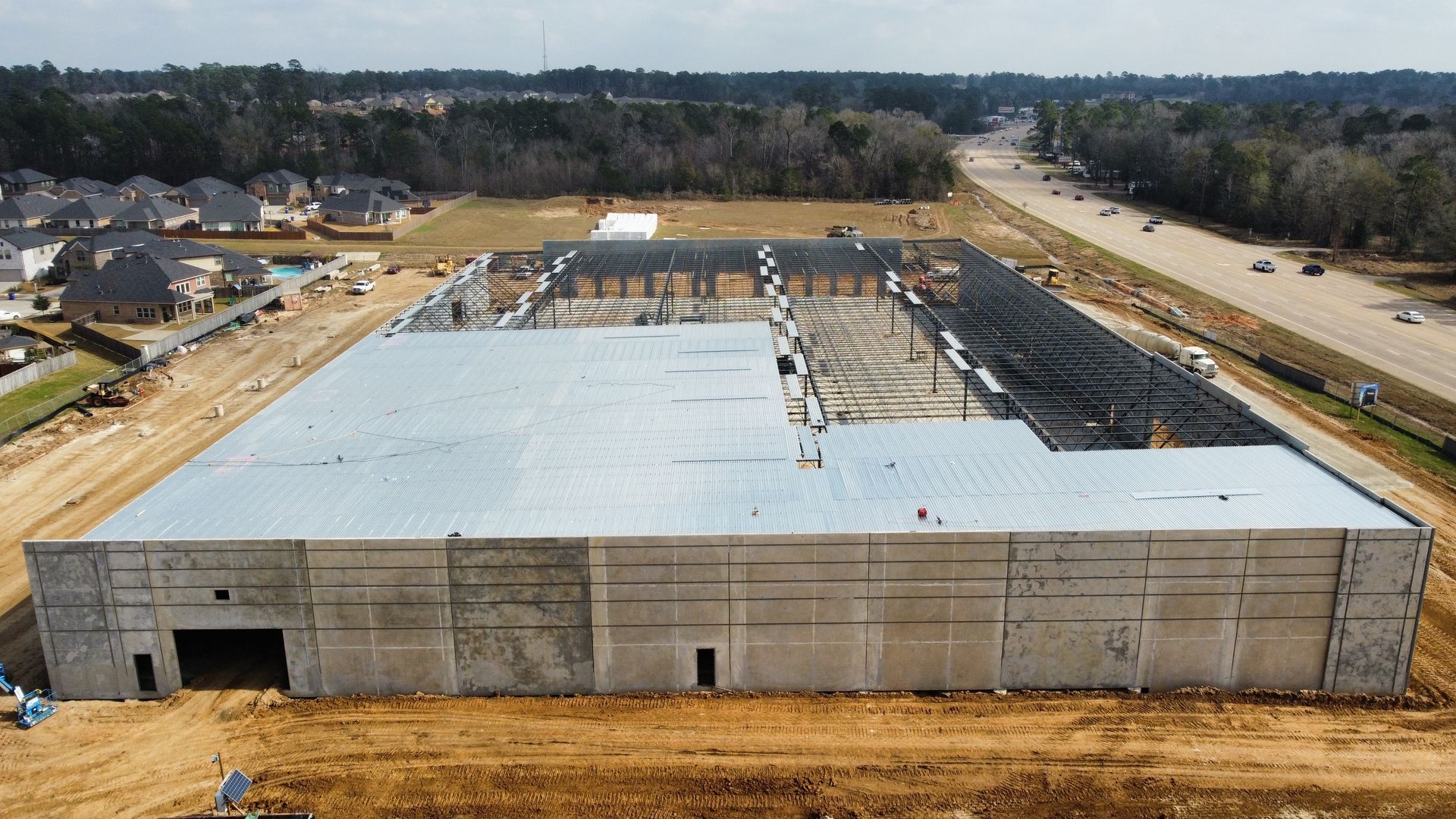 Construction site of a large commercial building; gray concrete walls and a partial roof. Dirt and highway visible.