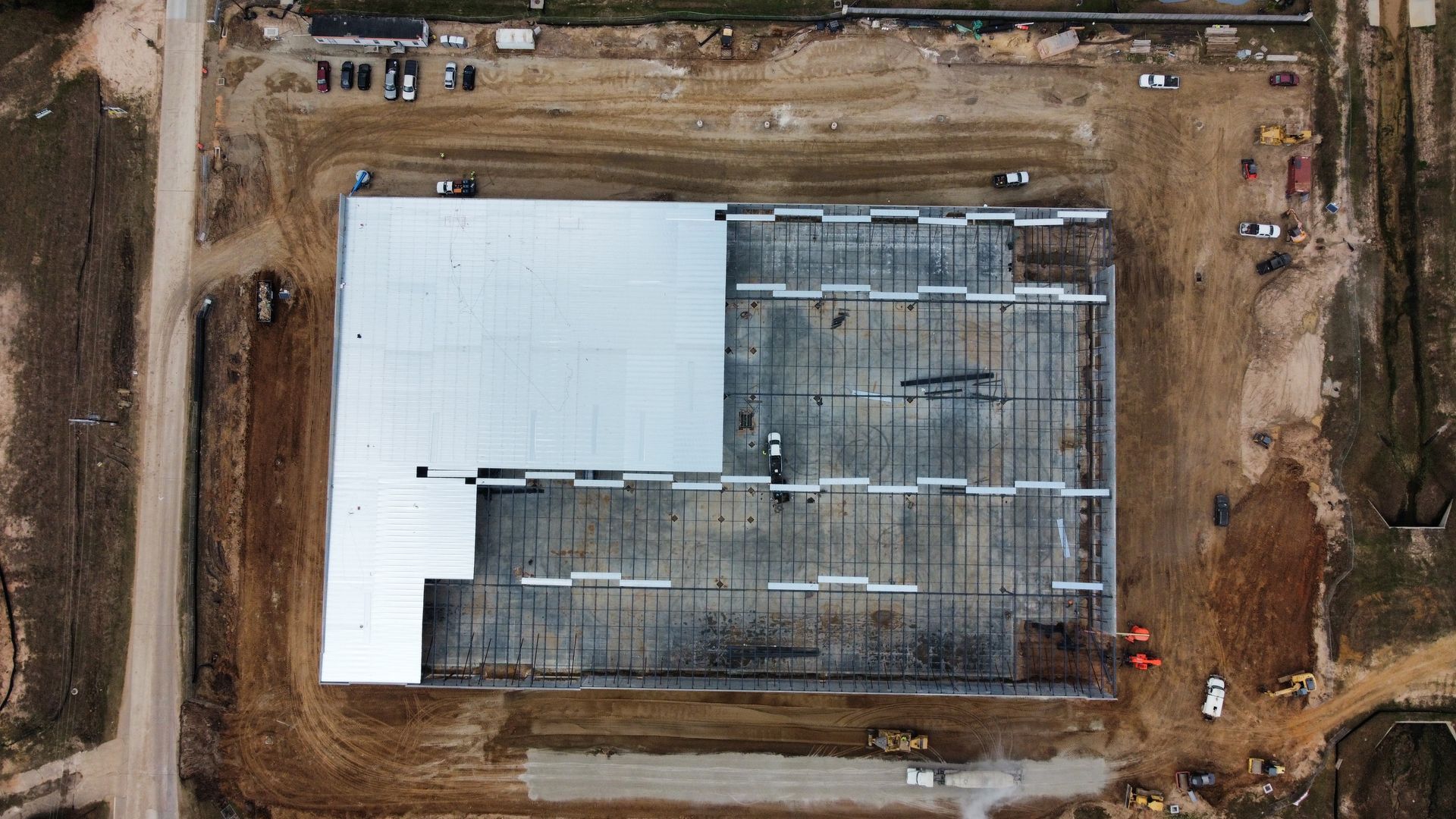 Aerial view of a partially constructed building with a silver and grey roof, surrounded by dirt and construction materials.