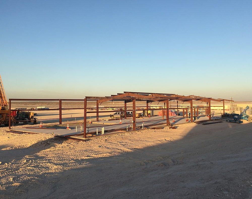 Steel framework of a building under construction on a dusty, flat landscape under a clear blue sky.
