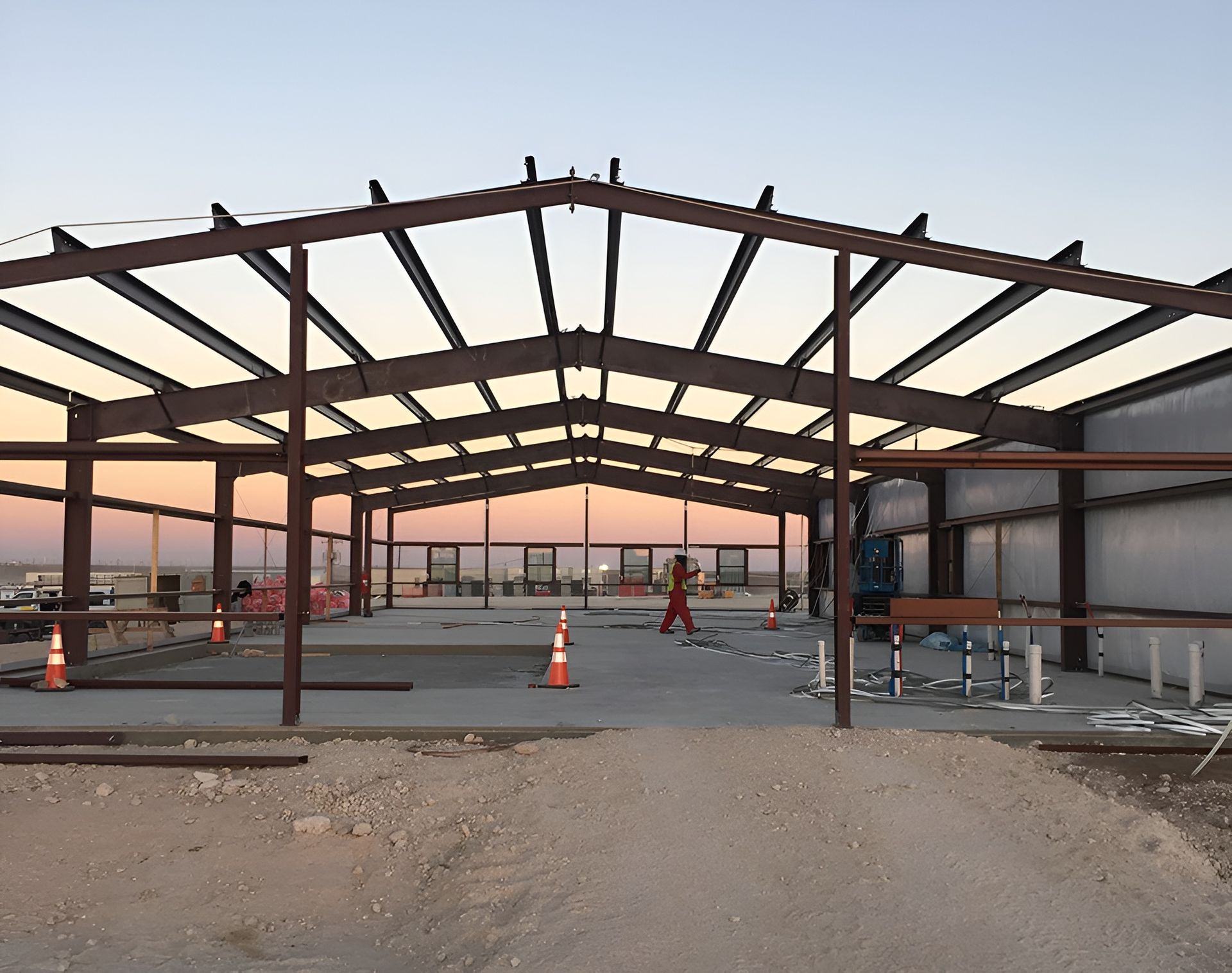 Steel framework of a building under construction, dusk sky in the background, construction worker walking, concrete floor.