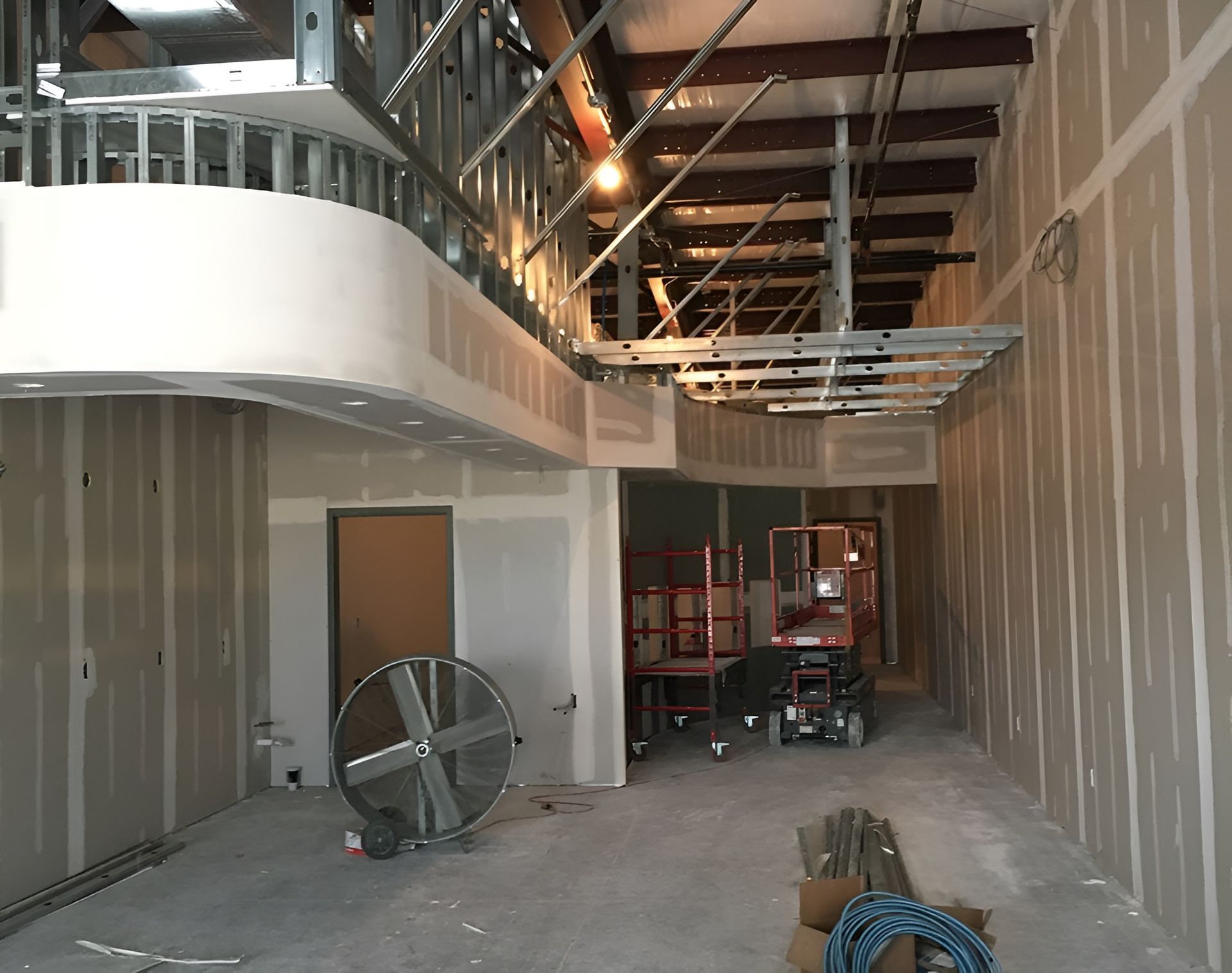 Interior construction scene with unfinished drywall, metal framing, and a large fan.