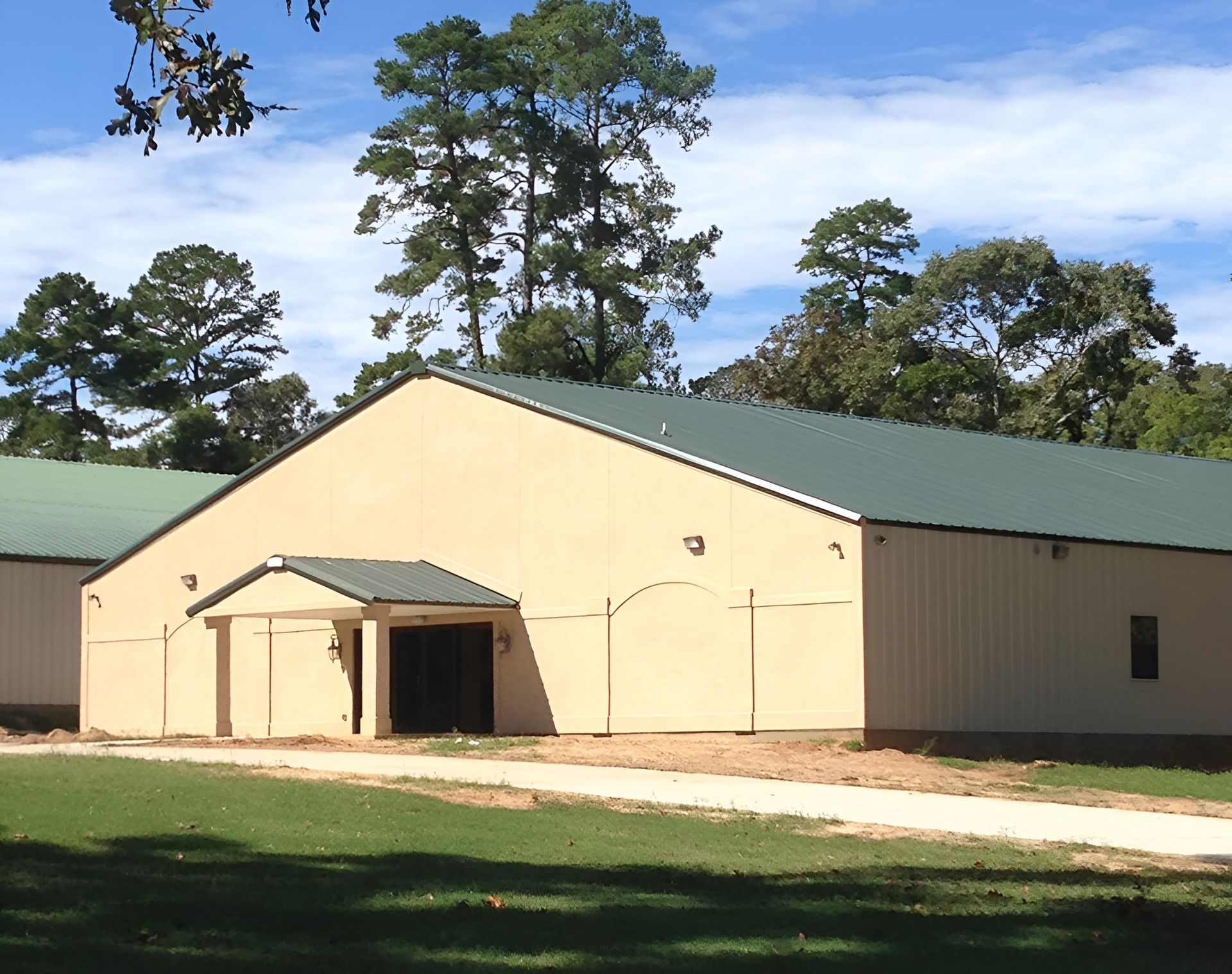 Beige metal building with green roof, trees in the background, a dirt path leading to the entrance.