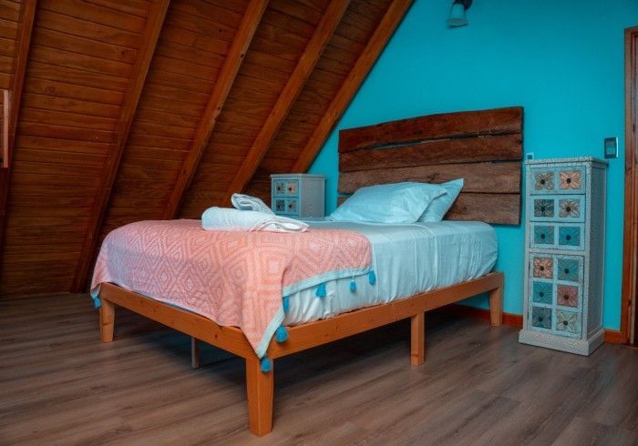 Bedroom with a wooden bed, headboard, and chest of drawers against a blue wall and slanted wood ceiling; a pink blanket and white linens.
