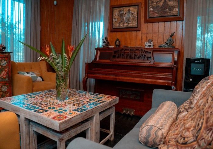 A living room with a vintage piano, colorful tile coffee table, yellow armchair, and blue couch. Wood paneling and artwork decorate the walls.