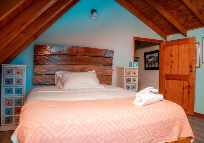Cozy bedroom with a bed, wooden headboard, and two small cabinets, under a slanted ceiling. A door leads to another room.