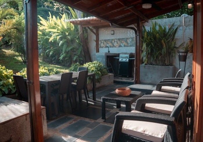 Outdoor patio with dining table, seating area with wicker furniture, and built-in grill. Lush green foliage in background.