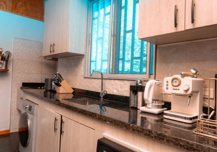 A well-lit kitchen featuring off-white cabinets, a dark countertop, and a window with a turquoise hue. Appliances include a coffee maker and kettle.