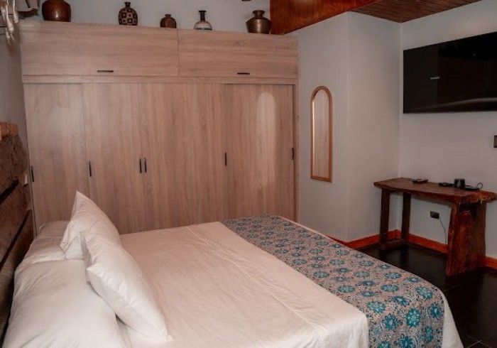 Bedroom with a bed, wooden wardrobe, TV, and a small wooden table against a white wall. Decorative jars sit atop the wardrobe.