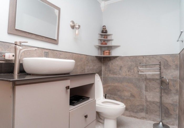 A modern bathroom featuring a white sink, toilet, and grey tiled walls. A small shelf displays decorative items.