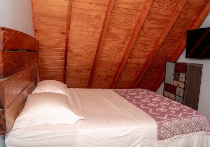 Bedroom with a bed under a sloped wood ceiling. The bed has white linens and a patterned pink throw; a wood headboard is on the left, and a cabinet with a TV are on the right.