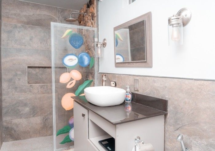 Bathroom with a glass shower door decorated with fruit, a vessel sink, and a small mirror. The walls have stone-like tiles.