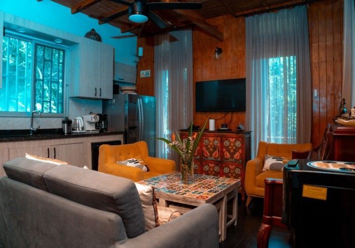 Living room with a gray sofa, yellow armchairs, and a colorful coffee table. Kitchen area in the background with stainless steel appliances.
