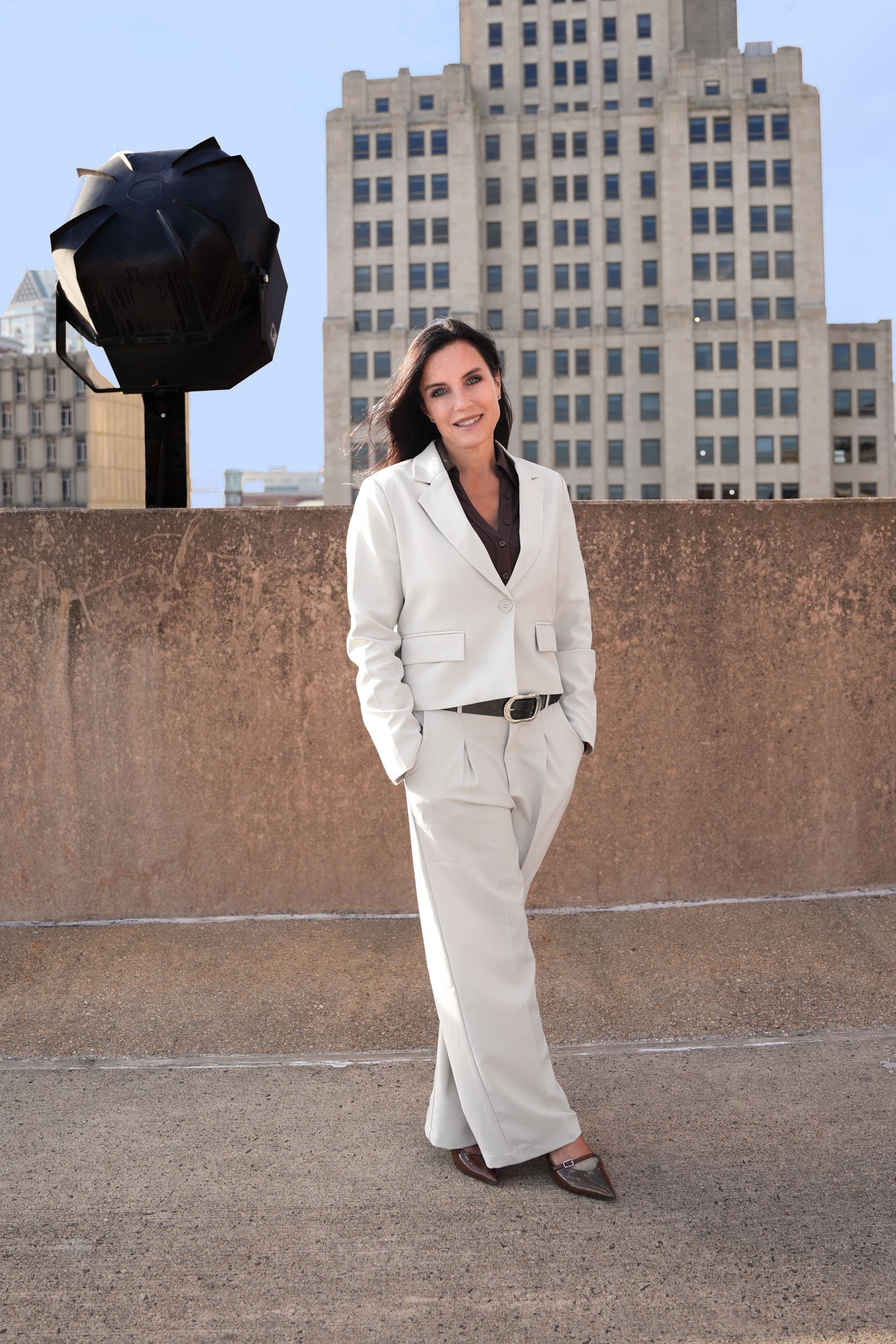 Woman in light suit with arms crossed, standing on a rooftop in front of a building.