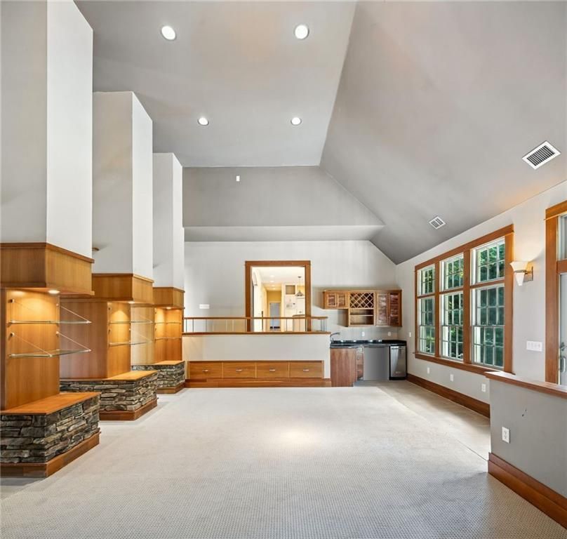 Spacious, open room with carpet, light walls, wooden accents, built-in shelving and bar area, and large windows.