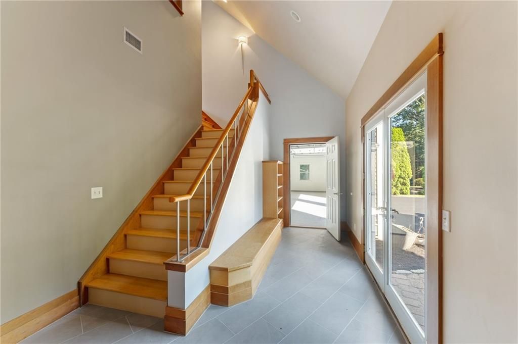 Interior hallway with wooden staircase, bench, and glass doors. Gray walls and flooring.