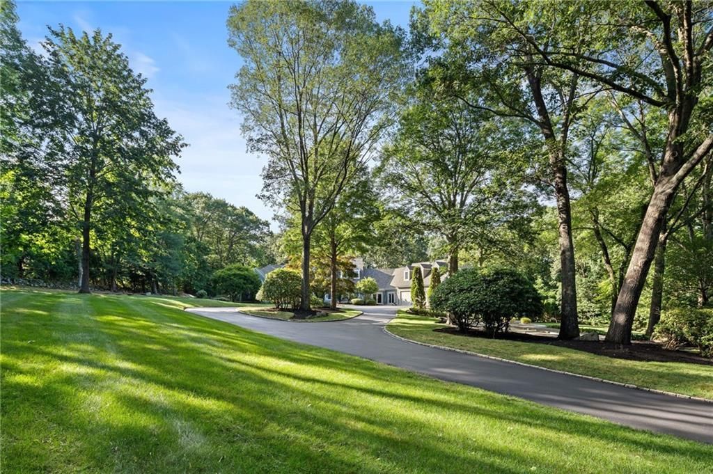 A long driveway leads to a house, surrounded by green grass and trees on a sunny day.