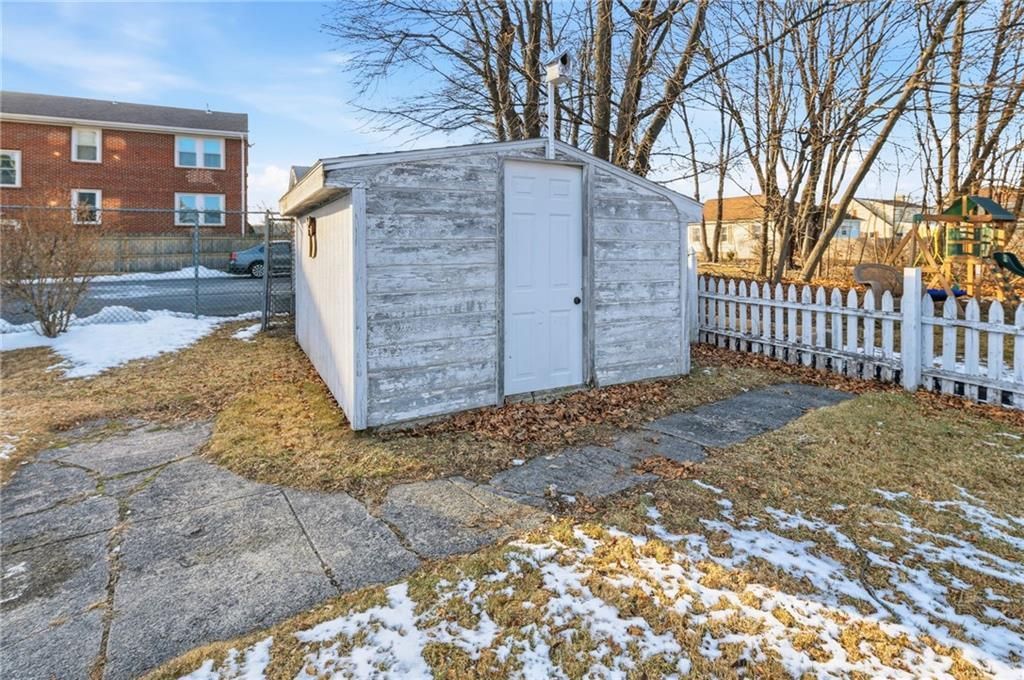 A small, weathered shed with a white door and a chimney. Next to a white picket fence, snow, and a cracked driveway.