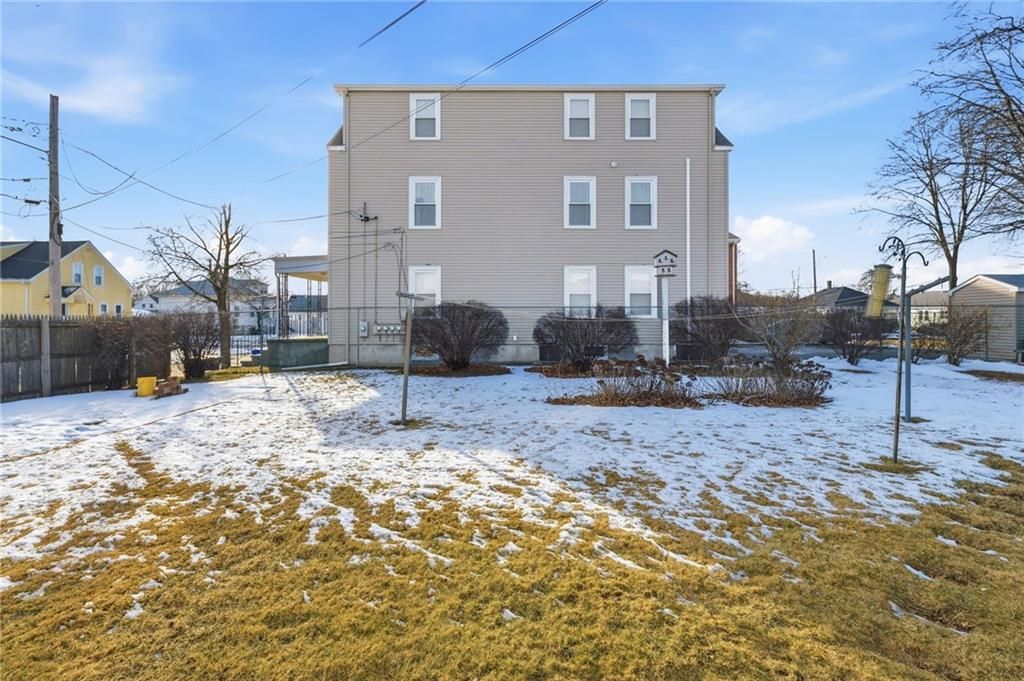 Rear view of a light beige three-story building with a snowy yard, leafless bushes, and a cloudy sky.