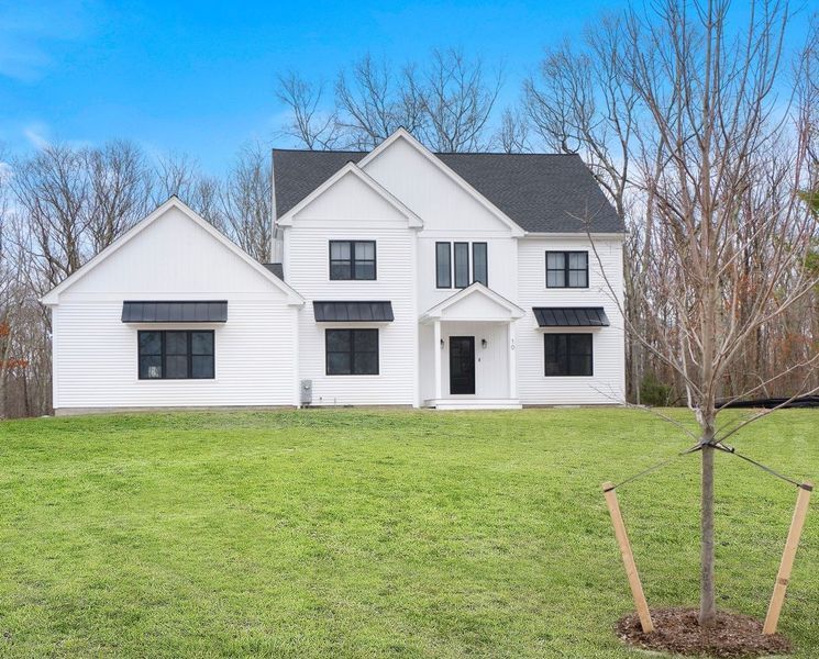 White two-story house with black trim and awnings, green lawn, tree in front, and trees in background under a blue sky.