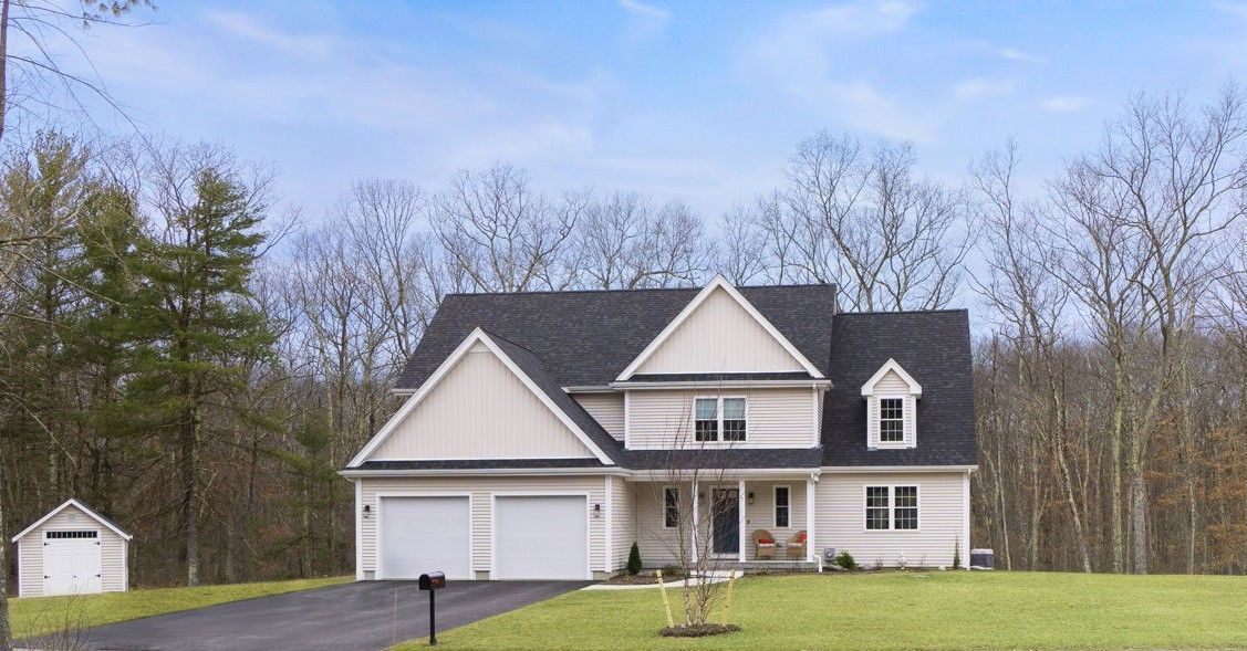 Two-story beige house with a dark roof and a small shed in a wooded setting.