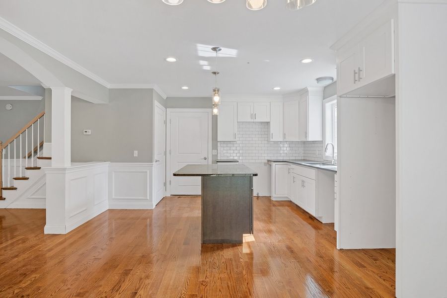 Open-concept kitchen with white cabinets, gray island, hardwood floors, and stairs in the background.
