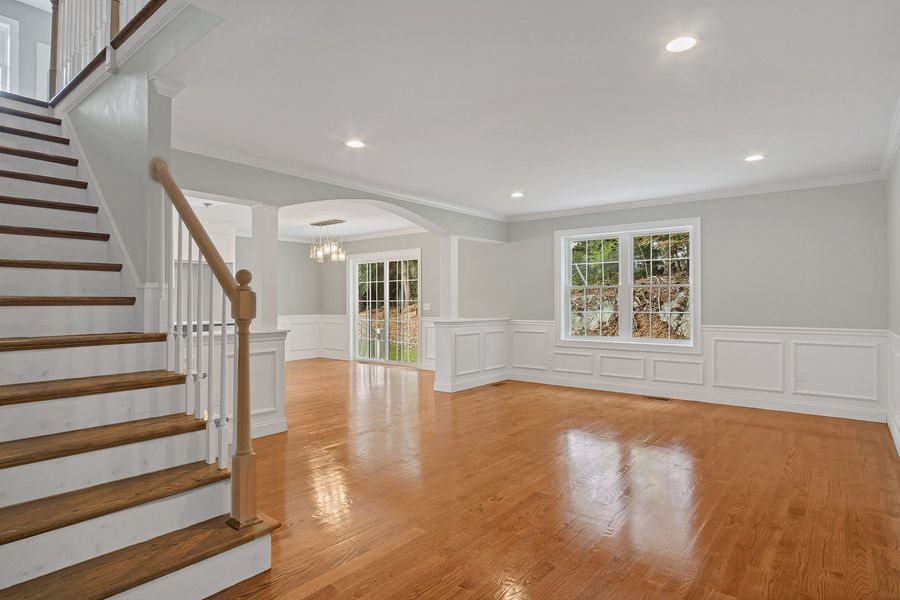 Empty living room with wood floors, staircase, and large windows.