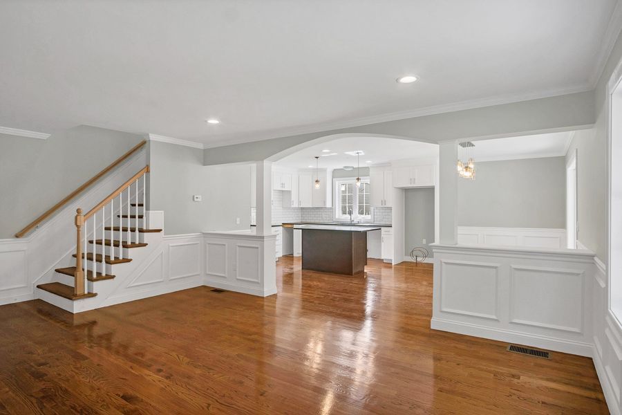 Empty living room with wood floors, staircase, and open kitchen visible through an archway.