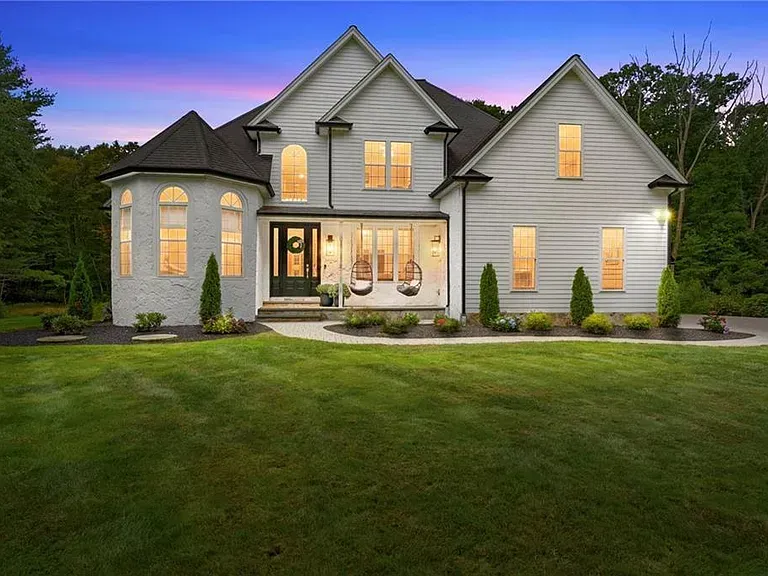 White two-story house with black trim, porch with hanging chairs, and a large front lawn, under a twilight sky.