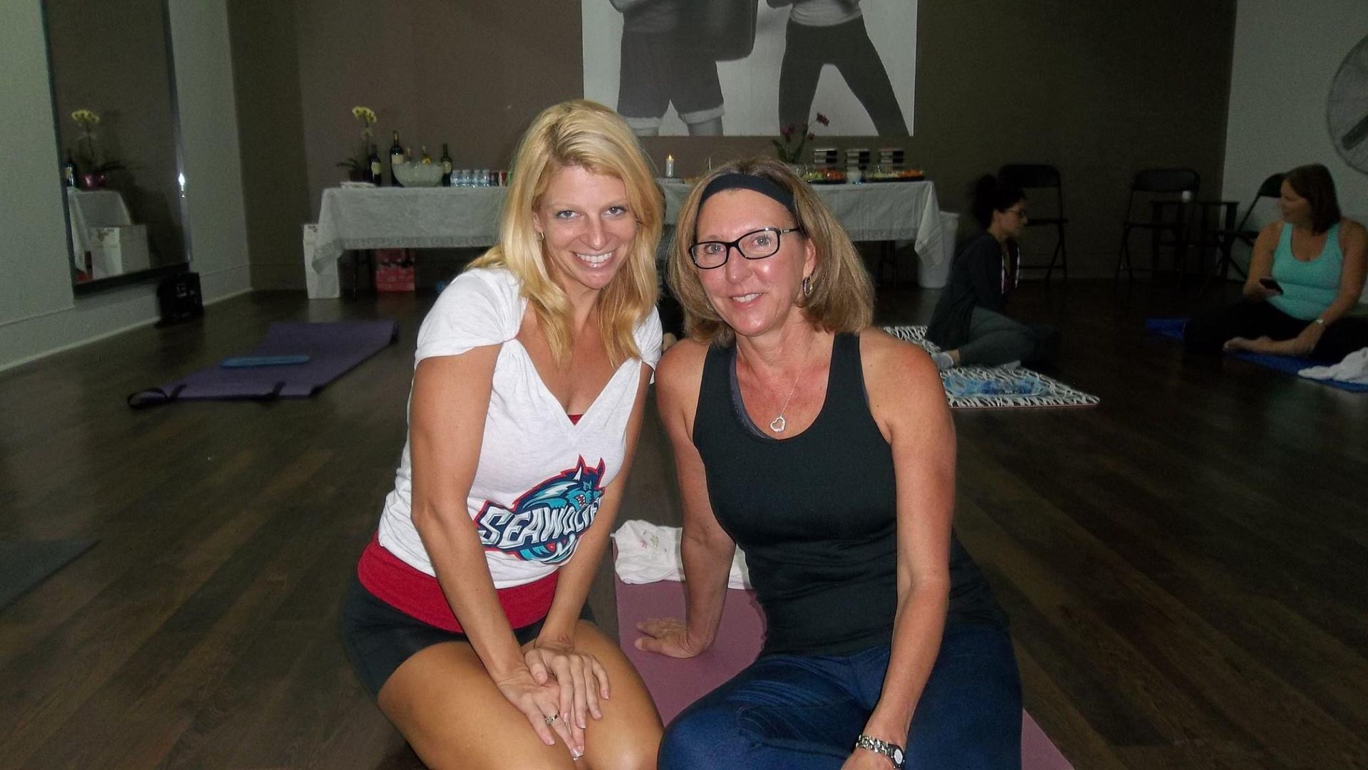 Two women are posing for a picture while sitting on a yoga mat