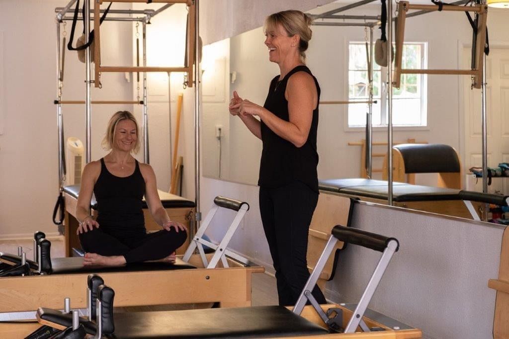 A woman is standing next to a woman sitting on a pilates machine in a gym.