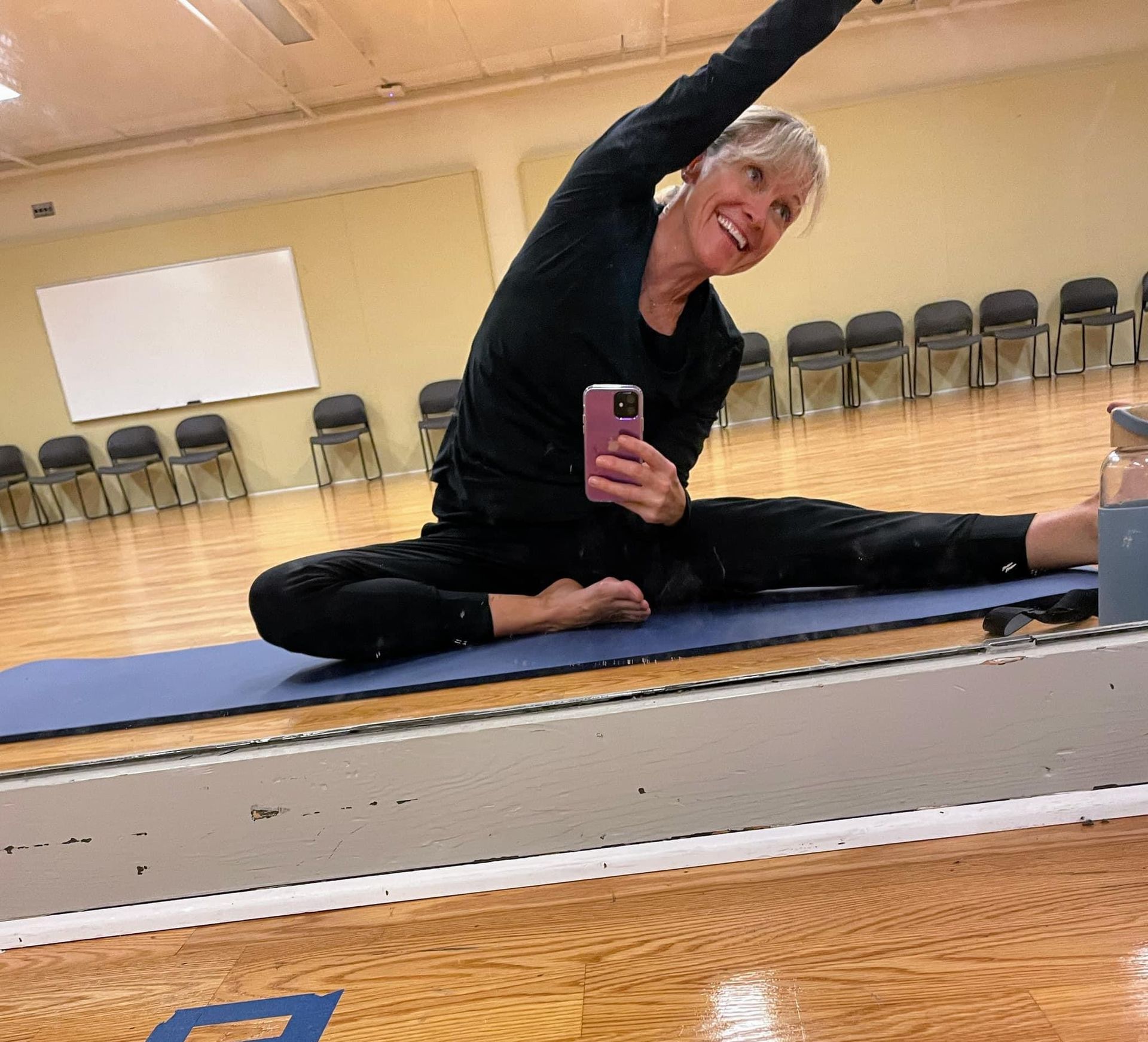 A woman is taking a selfie in front of a mirror while sitting on a yoga mat
