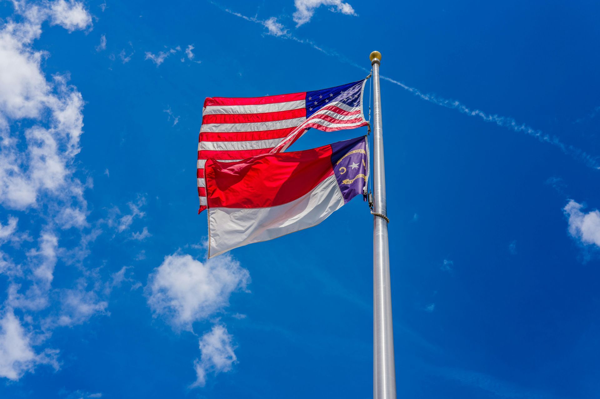 American flag and red and white flag on a pole against a blue sky with wispy clouds.