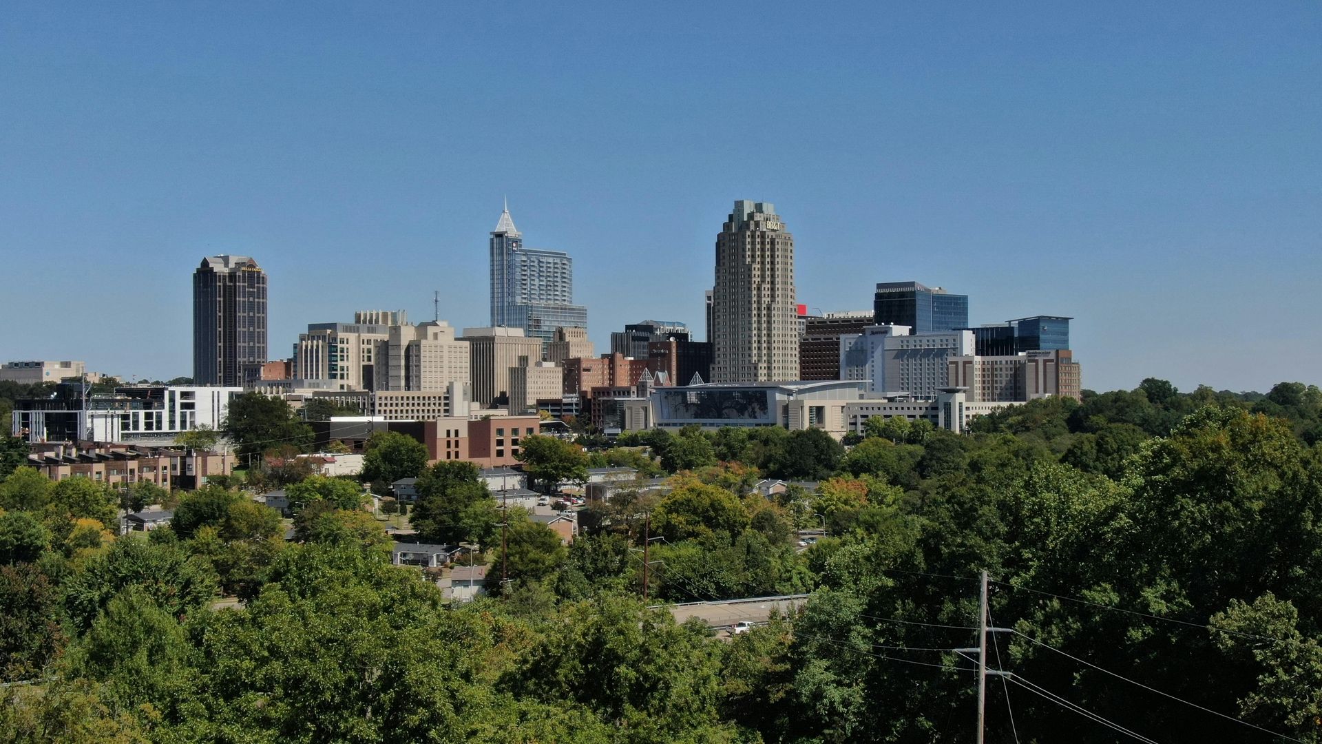 Skyline of a city with several tall buildings and a few smaller buildings, seen above a line of green trees under a clear, blue sky.