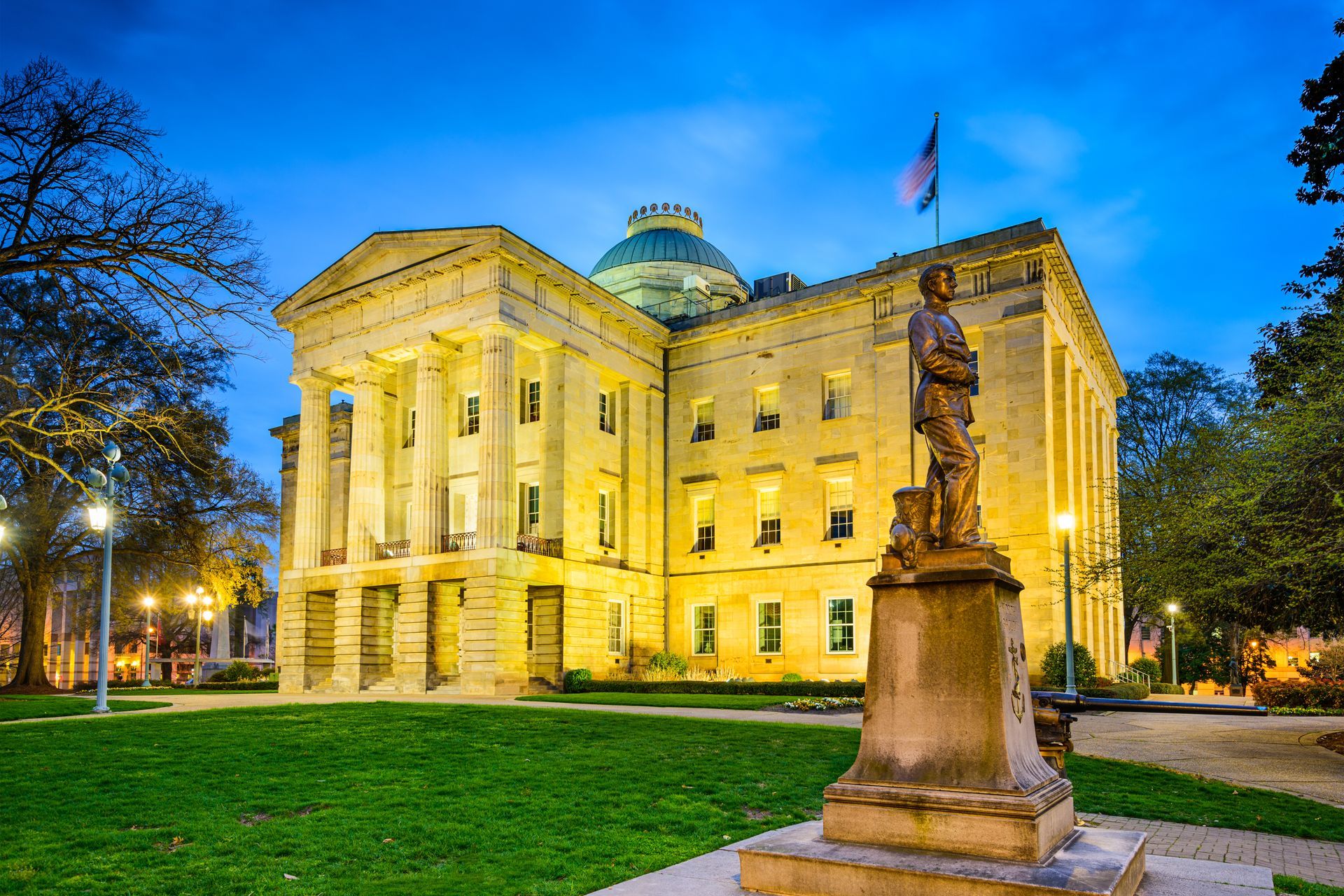 The North Carolina State Capitol building at dusk, with a statue in the foreground. The building is lit with yellow lights and the sky is blue.