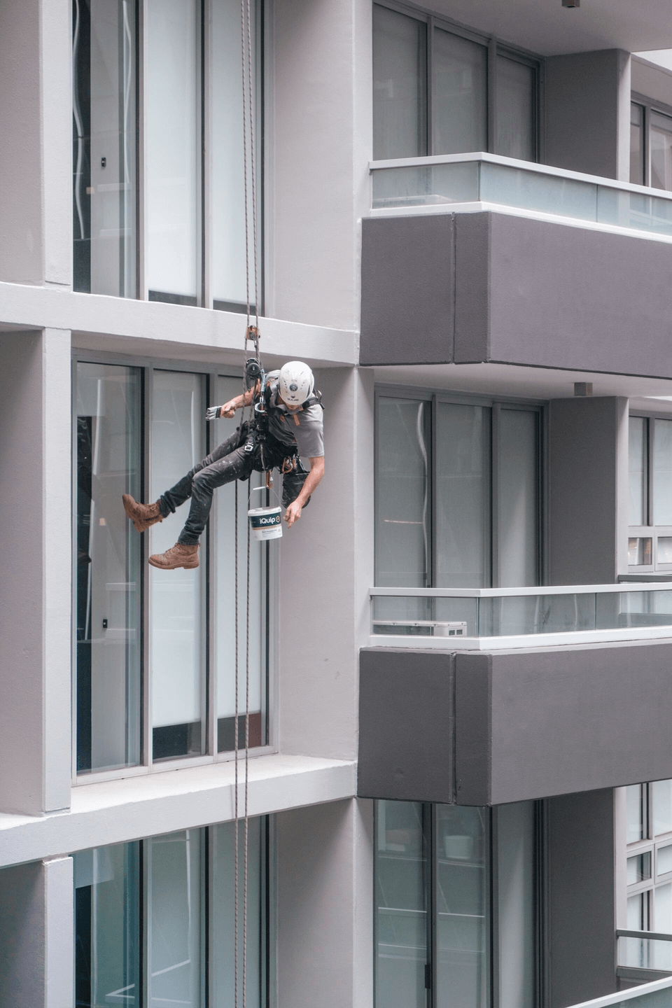 This is a picture of a guy who is suspended from a commercial building. He is hanging from a suspension rope. He is painting the outside of a commercial building. He is reaching for his paint bucket which is hanging from his waist.