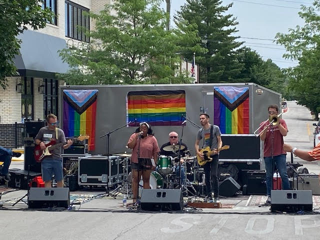 A group of people are playing instruments on a stage in front of a rainbow flag.
