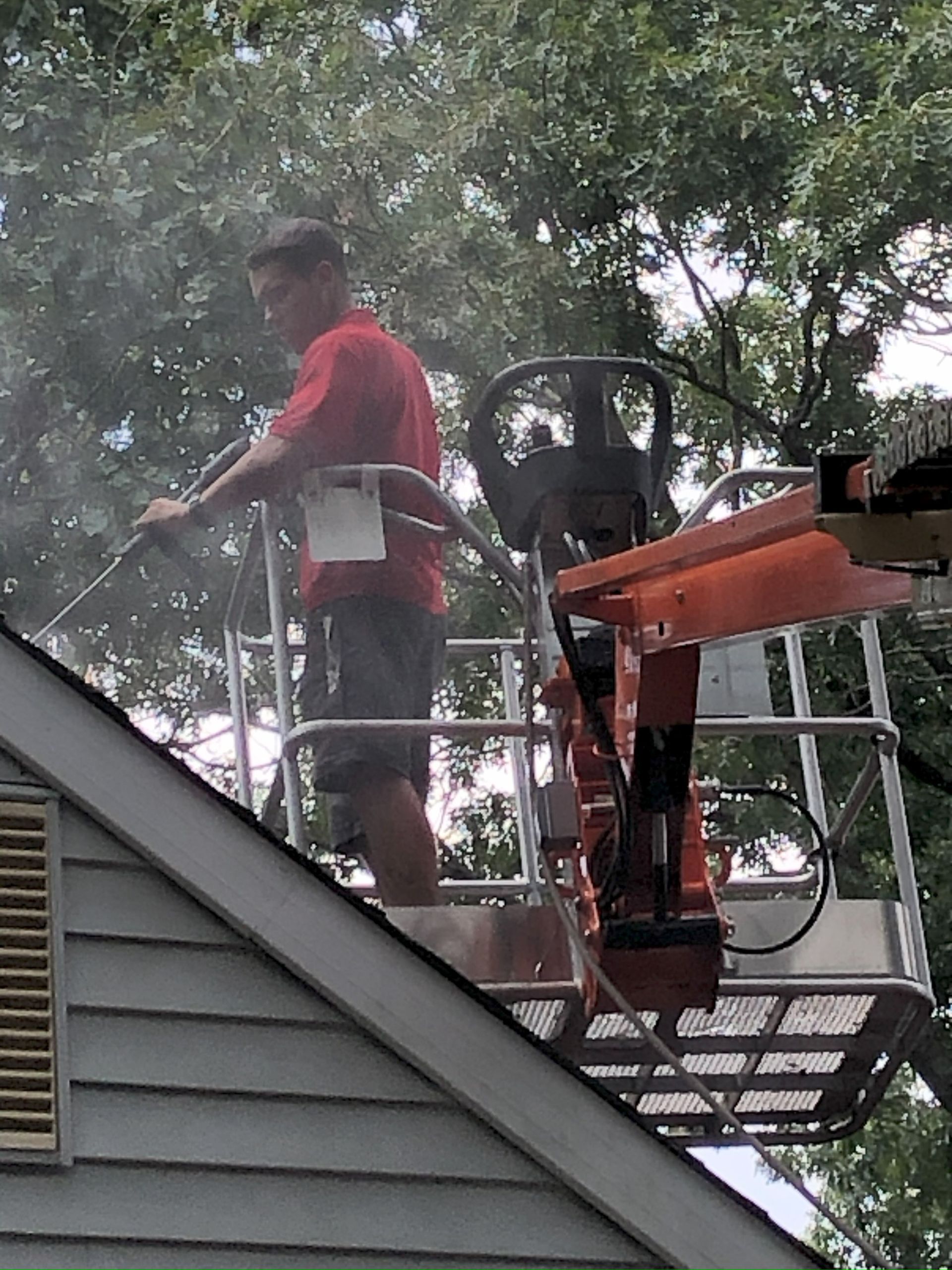 Man in a lift washing a house roof with a pressure washer. Gray siding, red shirt, and tree background.