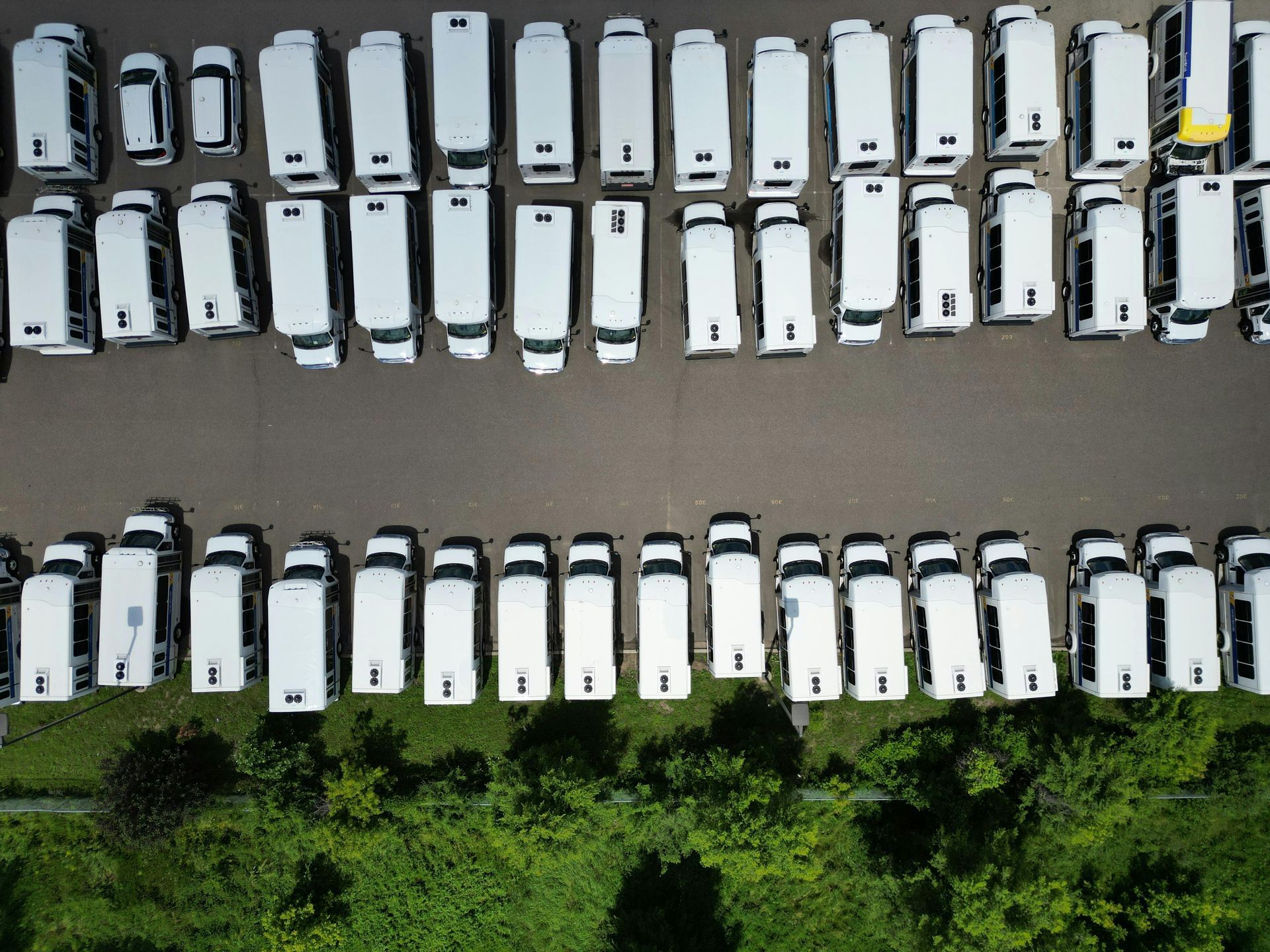 An overhead view shows rows of white vans parked in a lot, bordered by trees at the bottom.