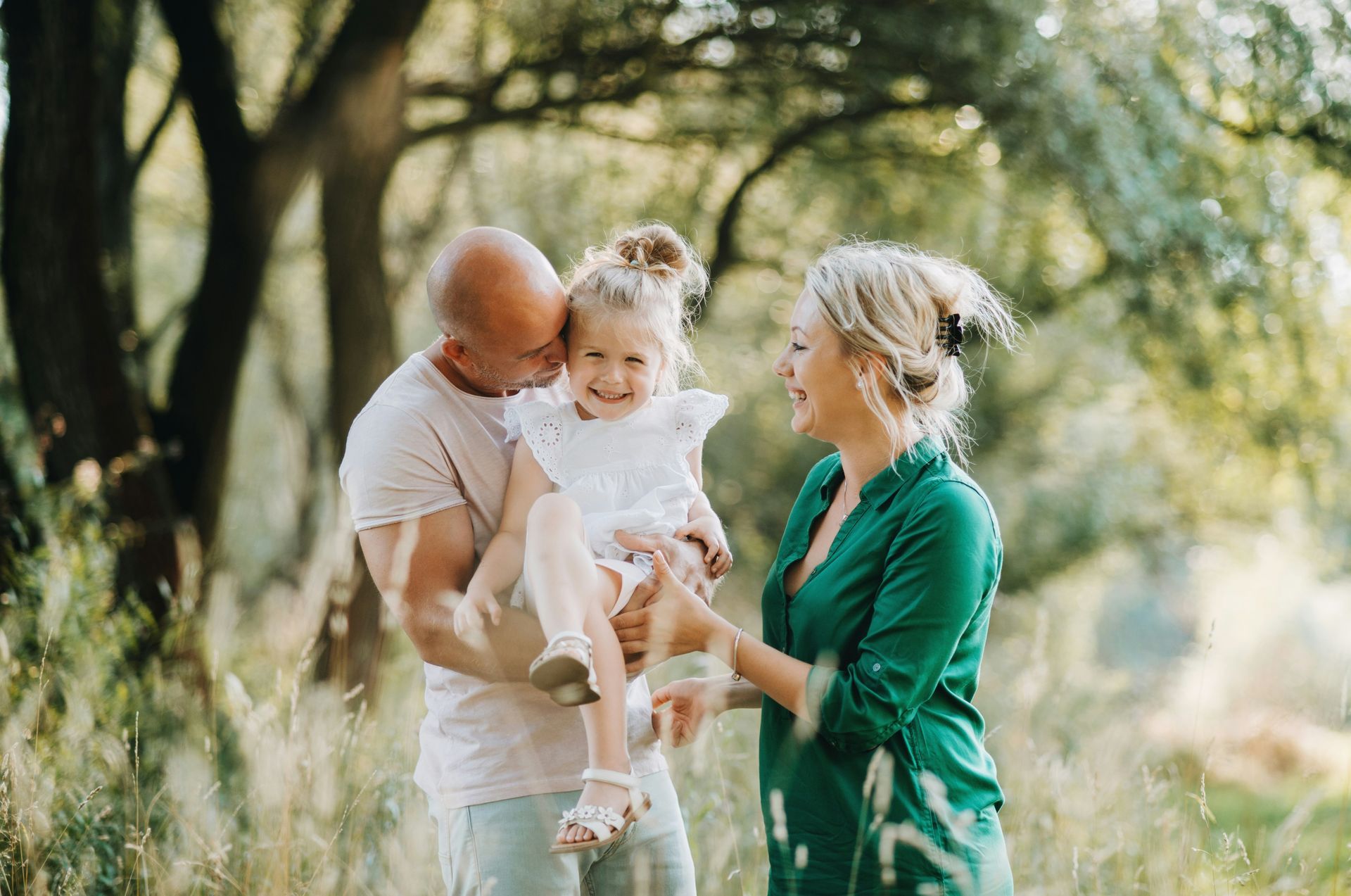 A family laughing together in a sunlit field, with an adult holding a young child while another adult reaches out.