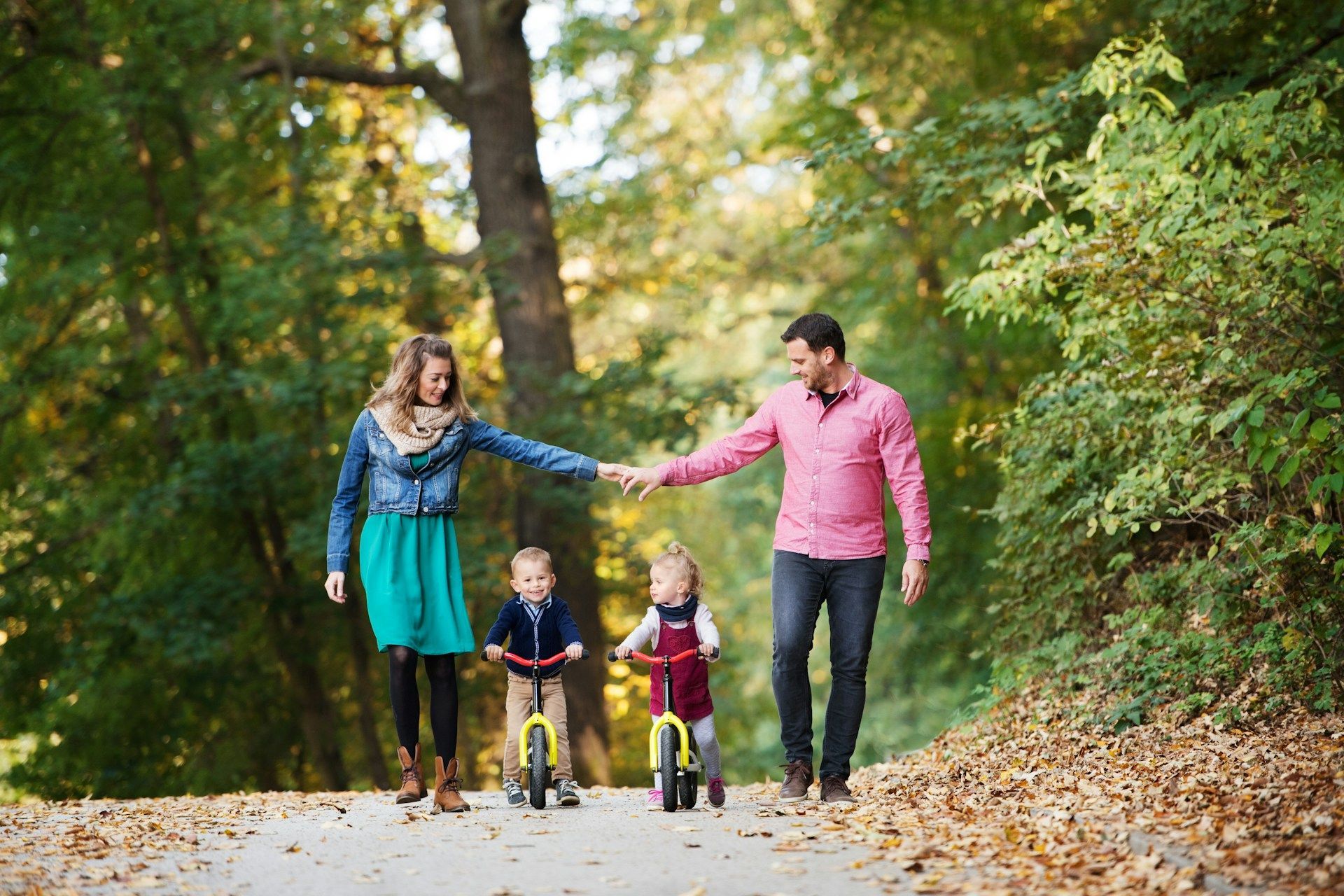 A family of four walking along a forest path in autumn, holding hands as two children ride balance bikes.