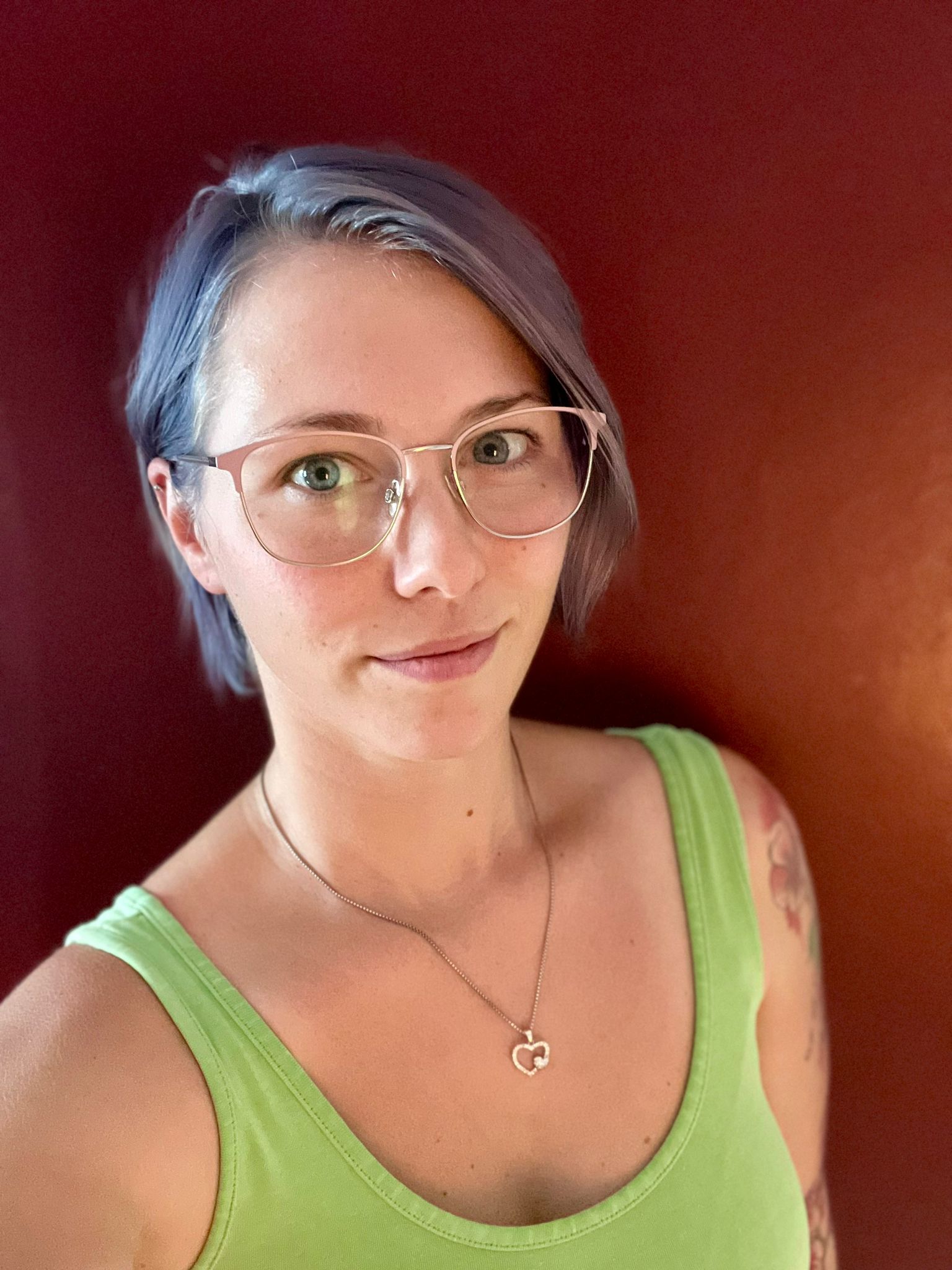 Eine Frau mit Brille und grünem Tanktop steht vor einer roten Wand.