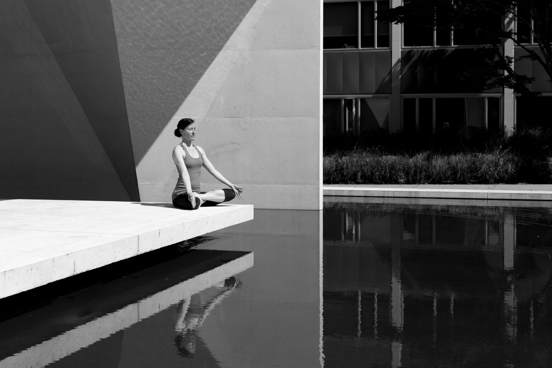A black and white photo of a woman meditating in front of a building and a pool.