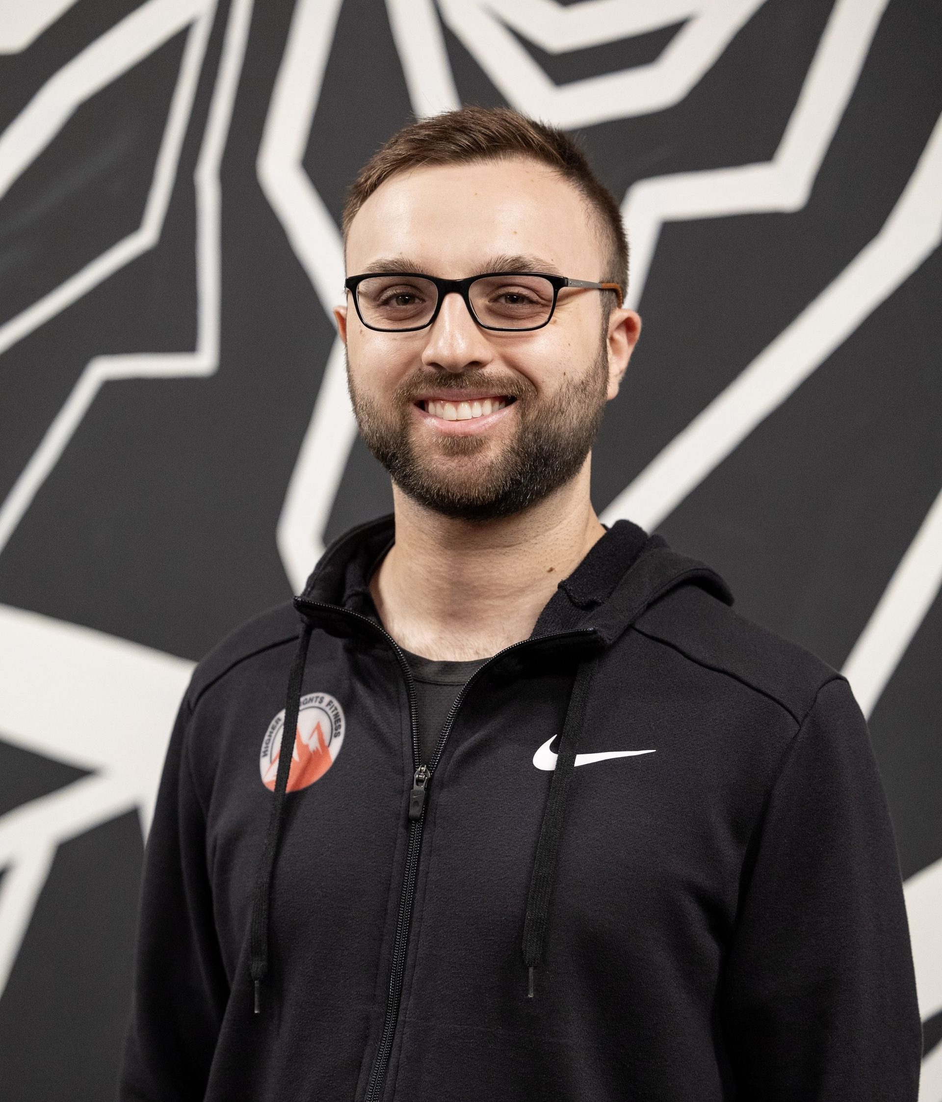 Man with glasses and beard smiles in front of green foliage, wearing a gray t-shirt with a mountain graphic.