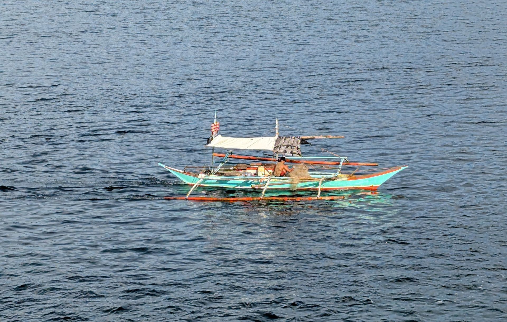 A small turquoise boat with outriggers floats on blue water. It has a white canopy, and a flag.